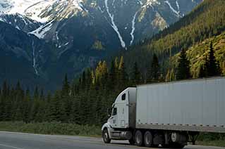 A white semi-truck driving on a road with tall pine trees and snow-capped mountains in the background under a clear sky.