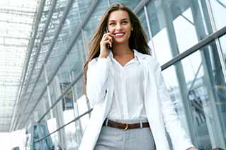 Woman in business attire talking on a smartphone, walking in a modern glass-walled office building.