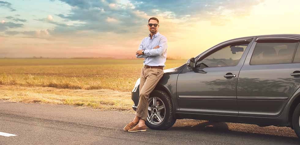 Man in sunglasses leaning against a gray car on a country road at sunset, with open fields and a colorful sky in the background.