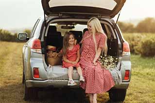 Mother and daughter sitting in the open trunk of a silver SUV in a field, smiling and holding flowers.