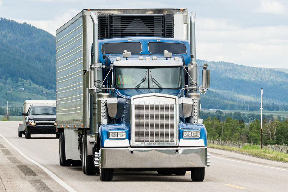 Front view of a blue semi-truck driving on a highway with mountains