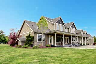 Large modern two-story house with beige siding, multiple gables, and a spacious front porch, surrounded by a well-maintained green lawn and small trees under a clear blue sky. Large modern two-story house with beige siding, multiple gables, and a spacious front porch, surrounded by a well-maintained green lawn and small trees under a clear blue sky.