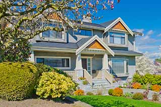 Two-story blue house with a porch, surrounded by green bushes and blooming trees under a clear blue sky. Two-story blue house with a porch, surrounded by green bushes and blooming trees under a clear blue sky.
